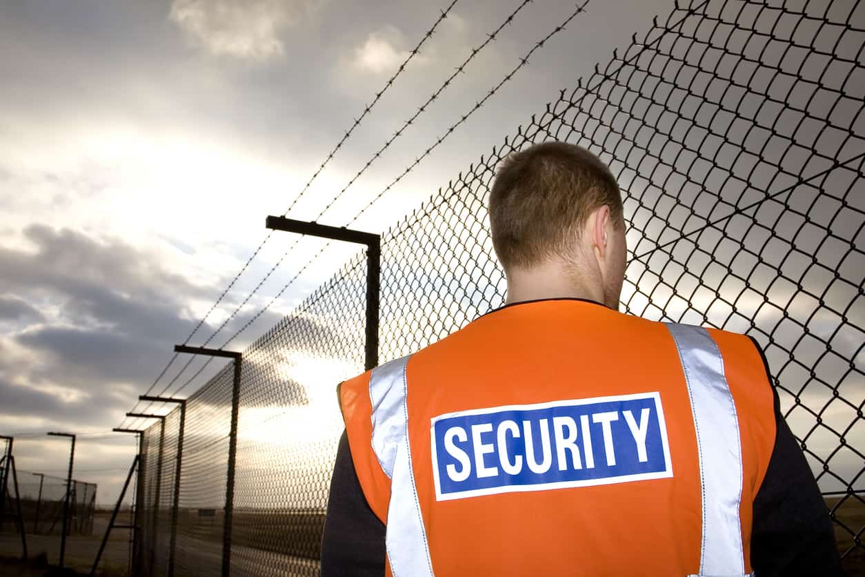 A security guard wearing an orang high vis vest with security written on the back patrolling a high wire perimeter fence.